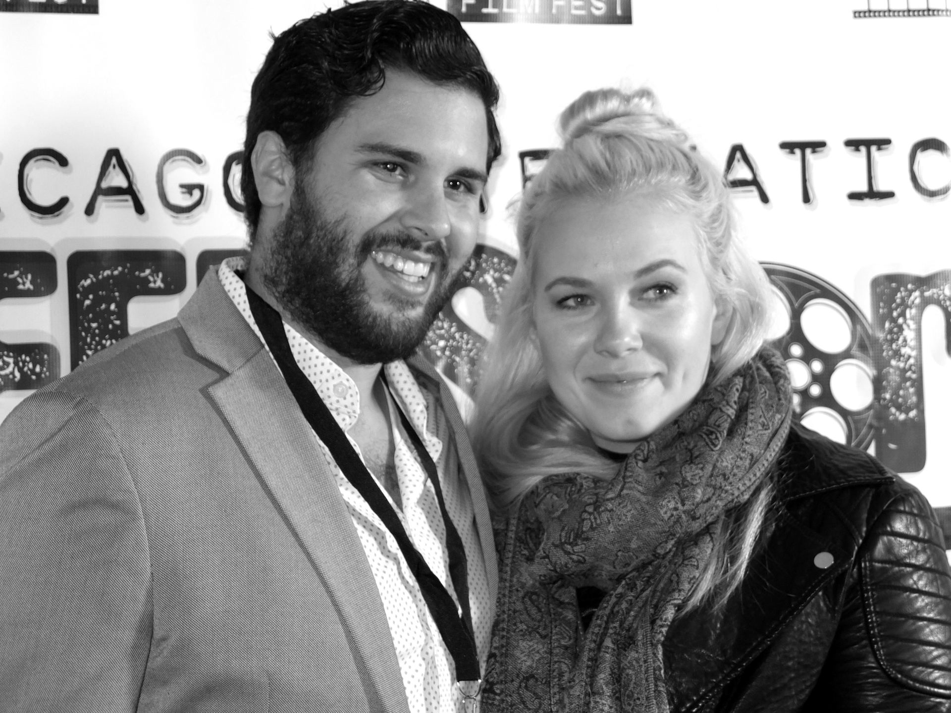 Man and woman at an event, smiling. Man with beard in suit jacket, woman with blonde updo in leather jacket.