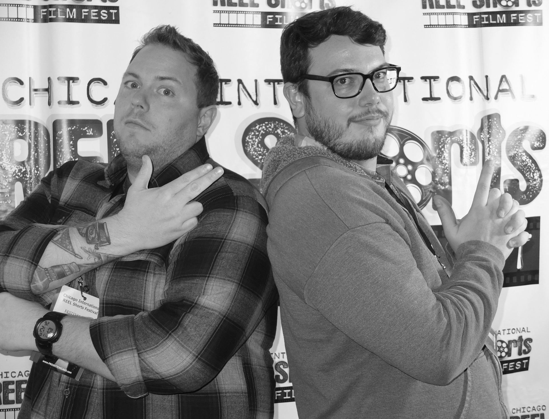 Two men posing at a film festival, making gun gestures. Black and white photo, backdrop visible.