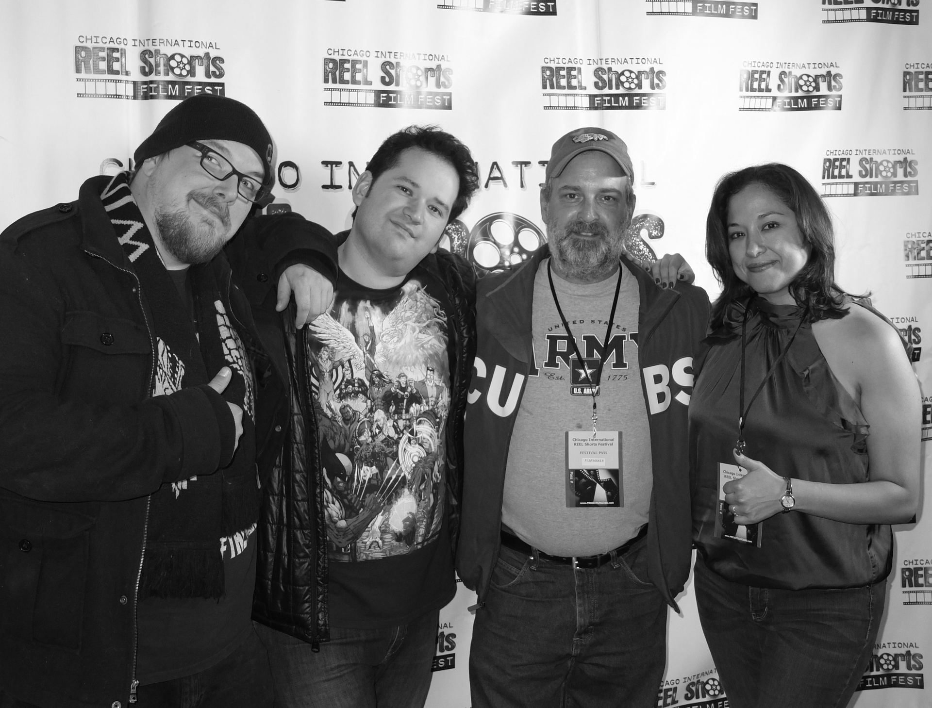 Four people pose at a film festival backdrop. Man in hat and glasses, man with arm around another, man with badge, and woman in halter top.