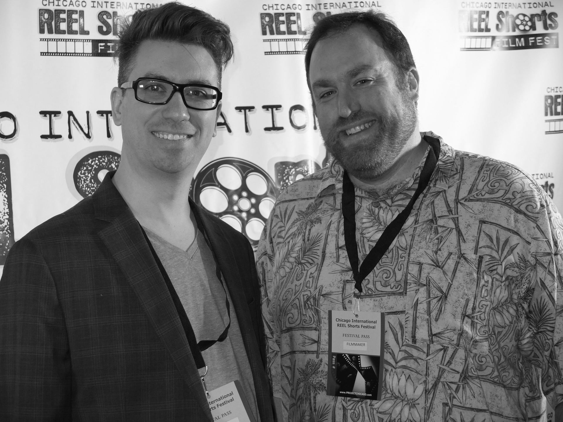 Two men smiling at a film festival. One wears glasses and a blazer, the other a tropical shirt.