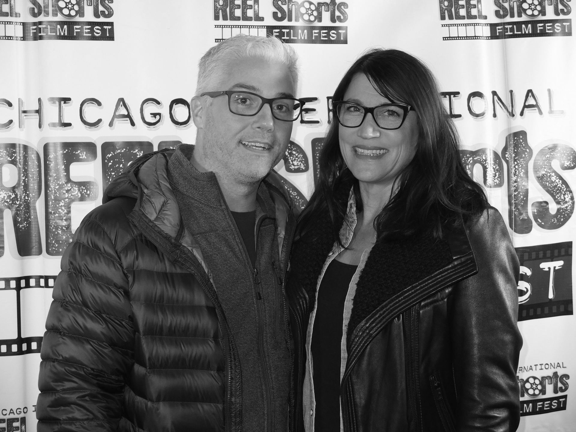 Man and woman at a film festival, smiling and wearing glasses, standing in front of a banner.