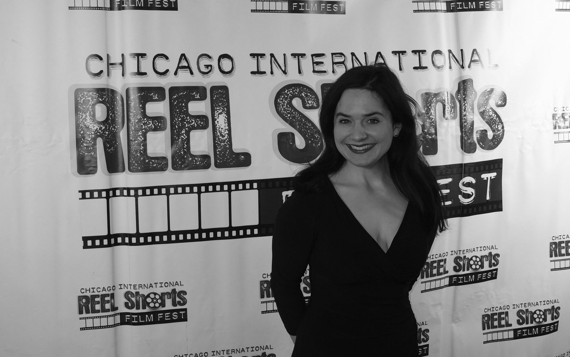 Woman at the Chicago International Reel Shorts Festival, smiling, wearing a black dress, posing in front of the festival backdrop.