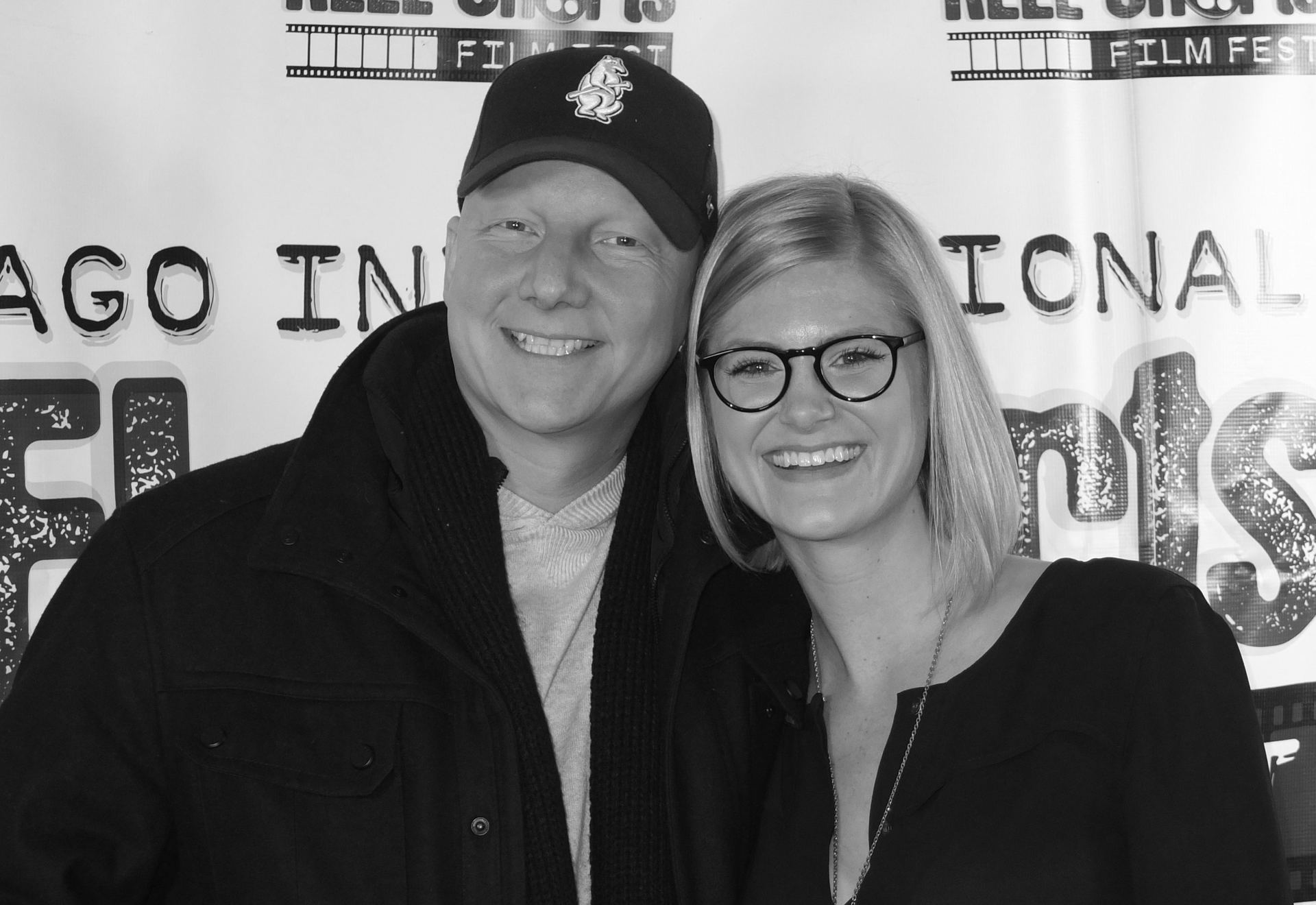 Man in cap and woman with glasses smile for a photo at a film festival.