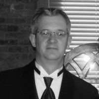 Man in a tuxedo and glasses; brick wall background, blinds to the right.
