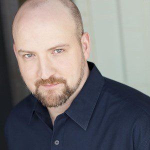 A bald man with a beard is wearing a blue shirt and looking at the camera.