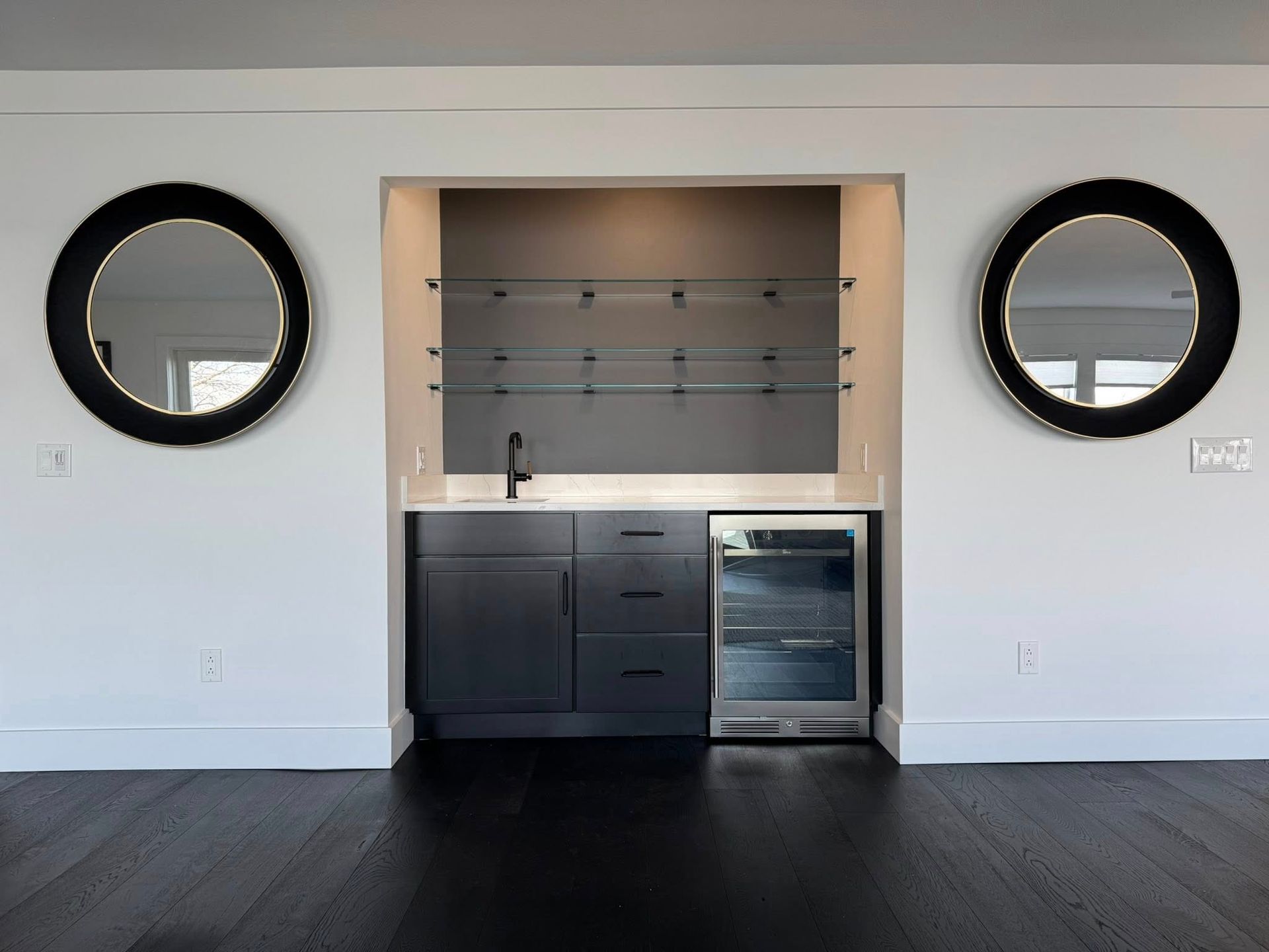 Built-in bar with gray cabinetry, a mini-fridge, shelving, and two circular mirrors on a white wall.