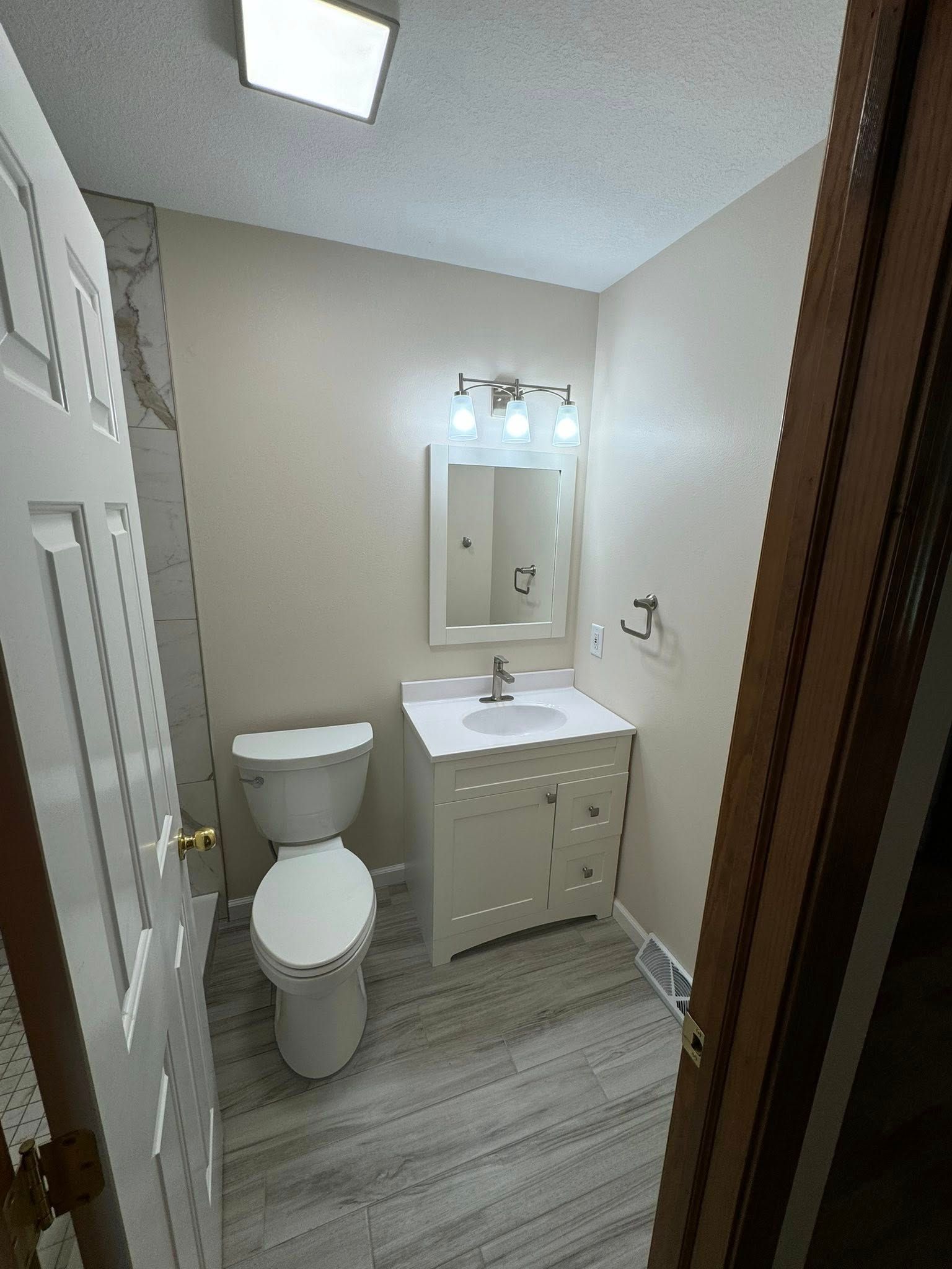 Small bathroom with white walls, vanity, and toilet, gray wood-look floor, and overhead light.