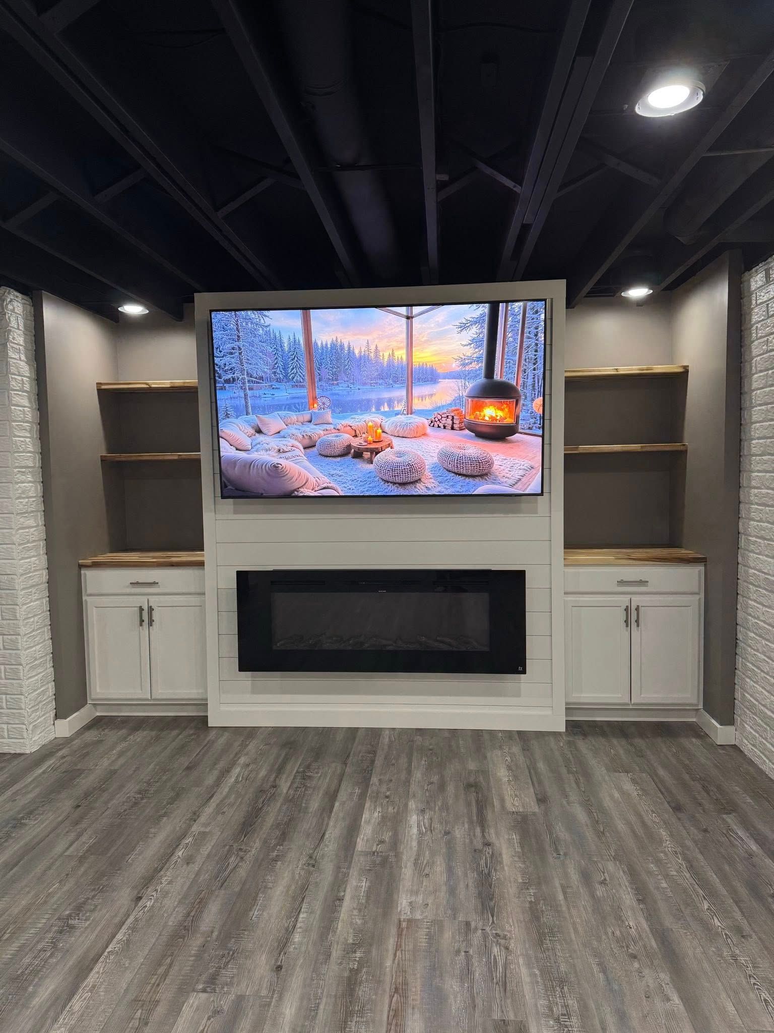 Living room with TV above a fireplace, flanked by cabinets and shelves. Dark ceiling, gray flooring.