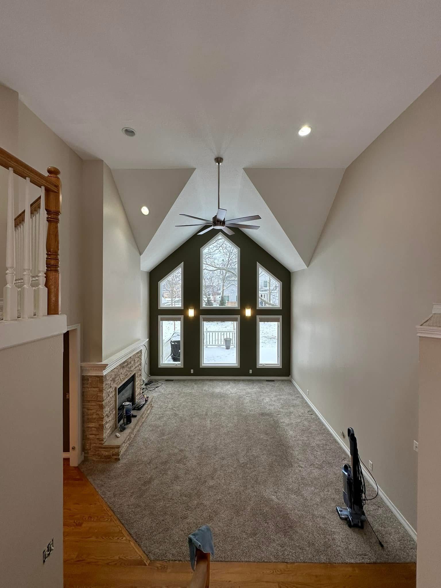 High-ceilinged living room with large windows, fireplace, and carpeted floor. A vacuum cleaner is in the corner.