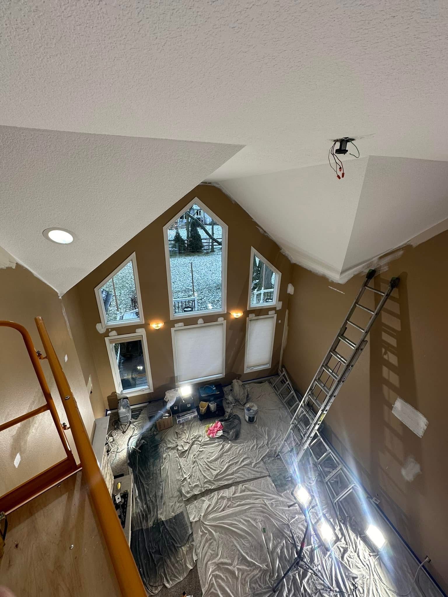 High-ceiling room under renovation with ladder, drop cloths, and workers visible near large windows.