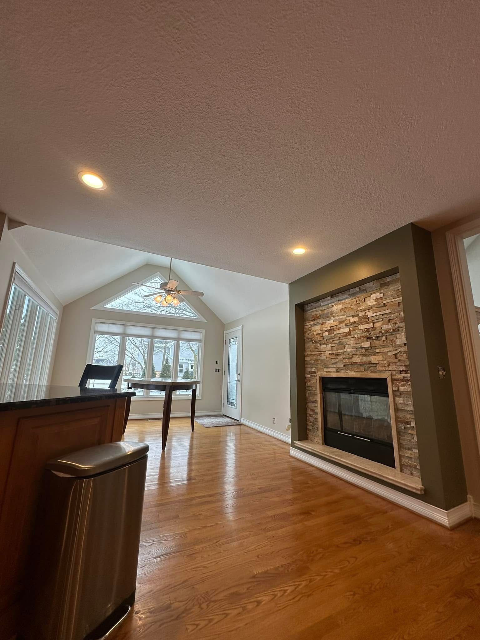 Living room with fireplace, hardwood floors, and vaulted ceiling.  A table sits near a triangular window.