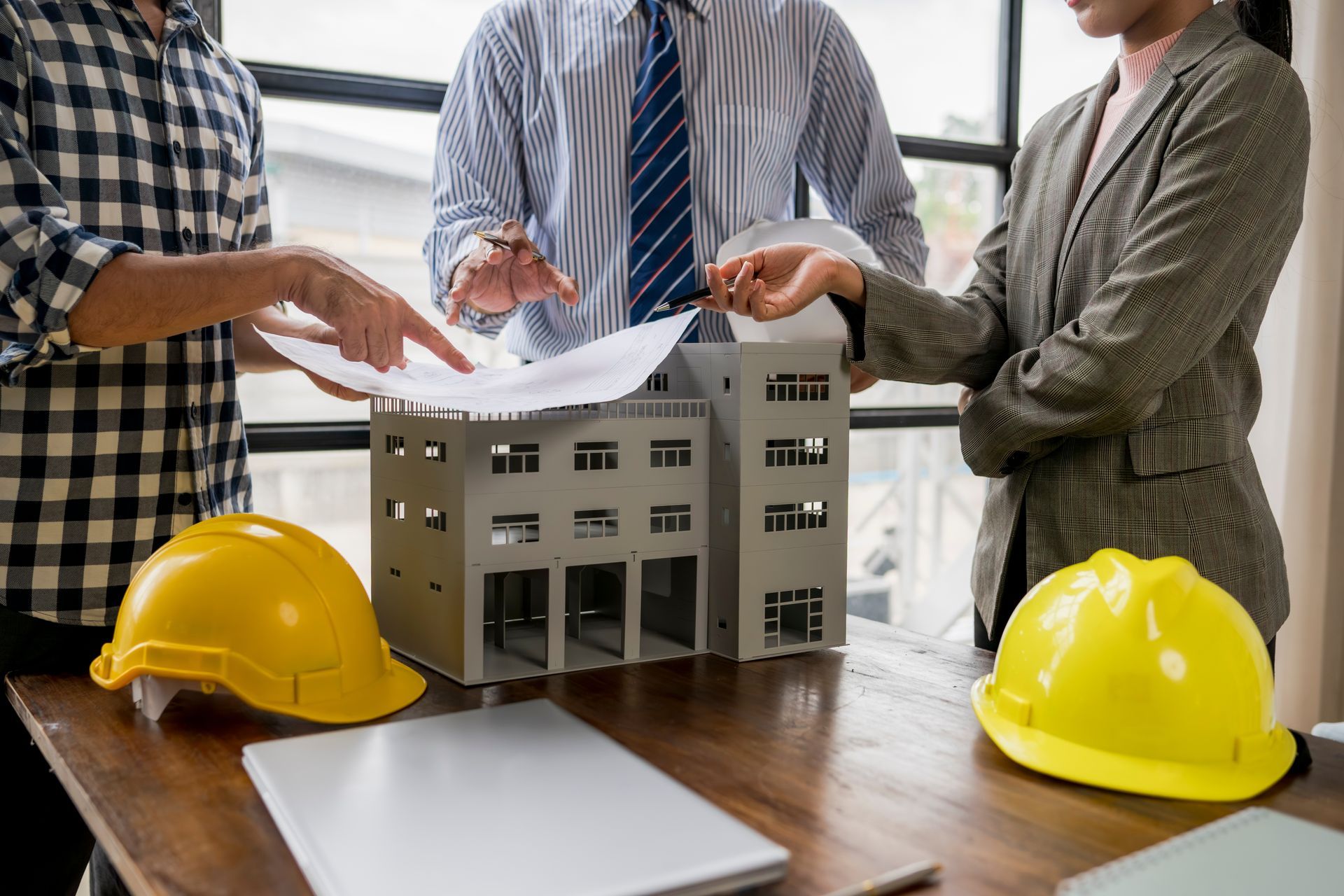 Three people in business attire reviewing blueprints over a building model with yellow hard hats on the table.