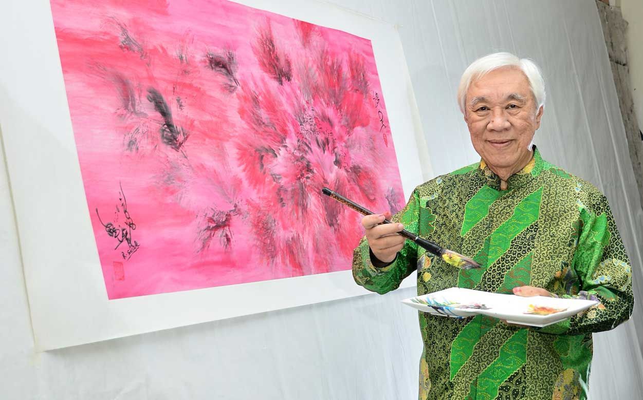 Artist with paintbrush and palette, painting abstract pink artwork. He wears a green patterned shirt, and smiles.