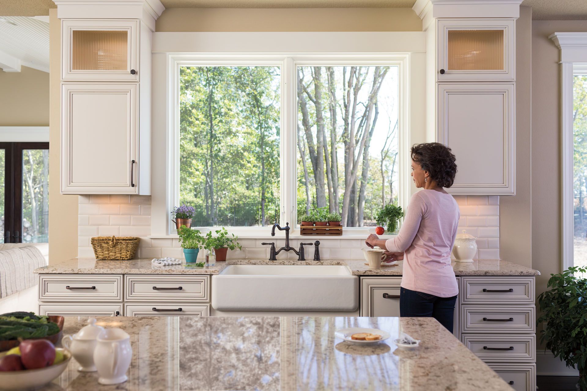 Woman in kitchen preparing food, looking out window at trees. White cabinets, granite counter.