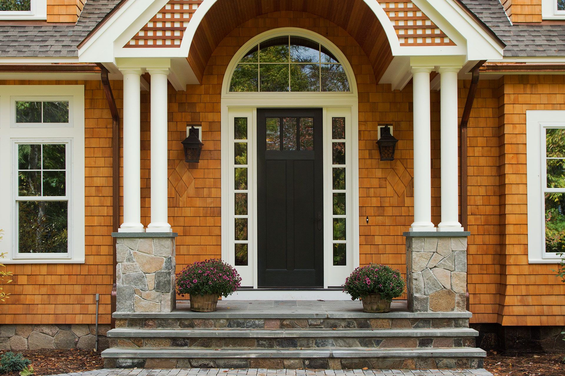 Brown-shingled house entrance with pillars, arched doorway, steps, and potted plants.