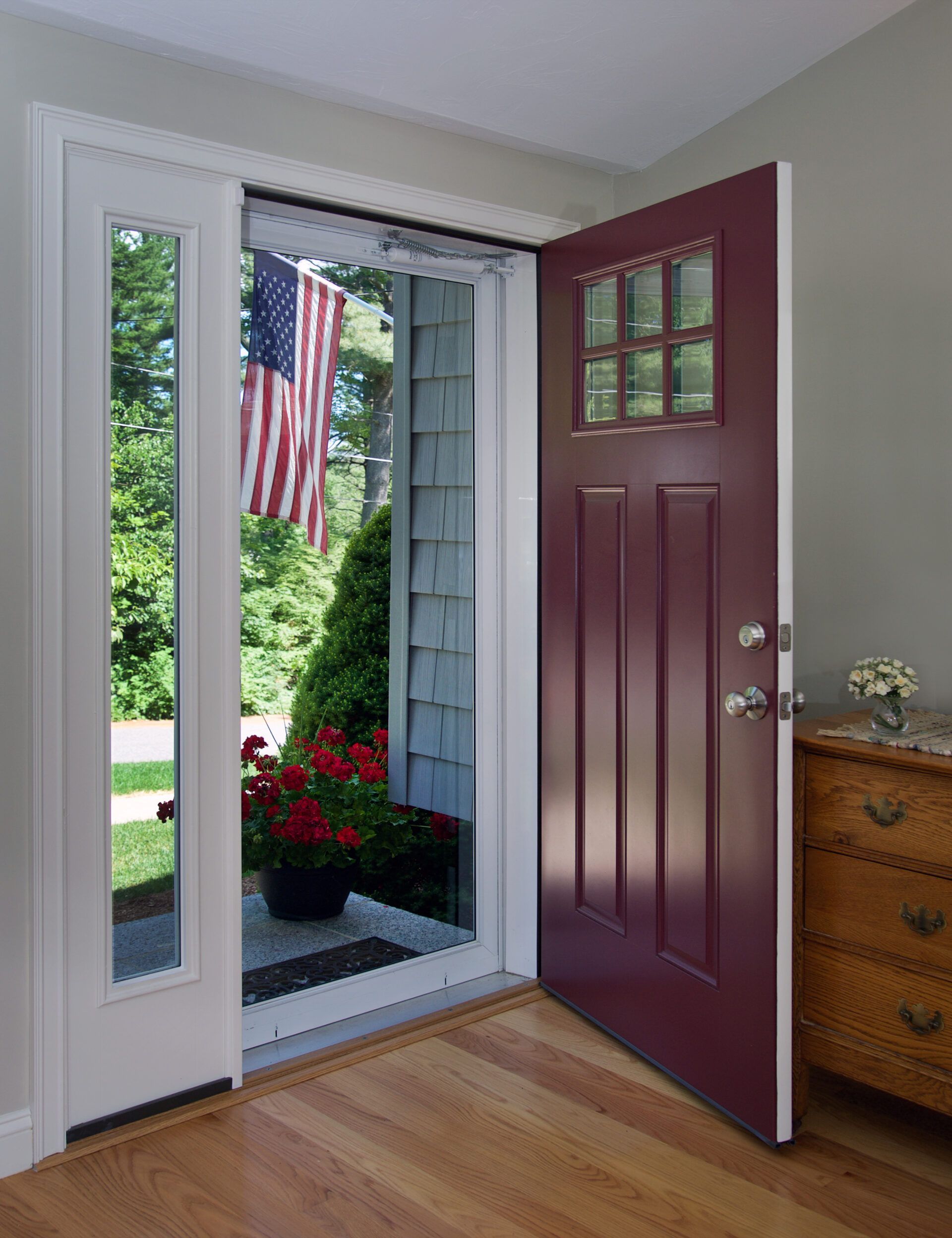 Red front door open to a view of a home, American flag visible. Interior has wood floors and a small dresser.