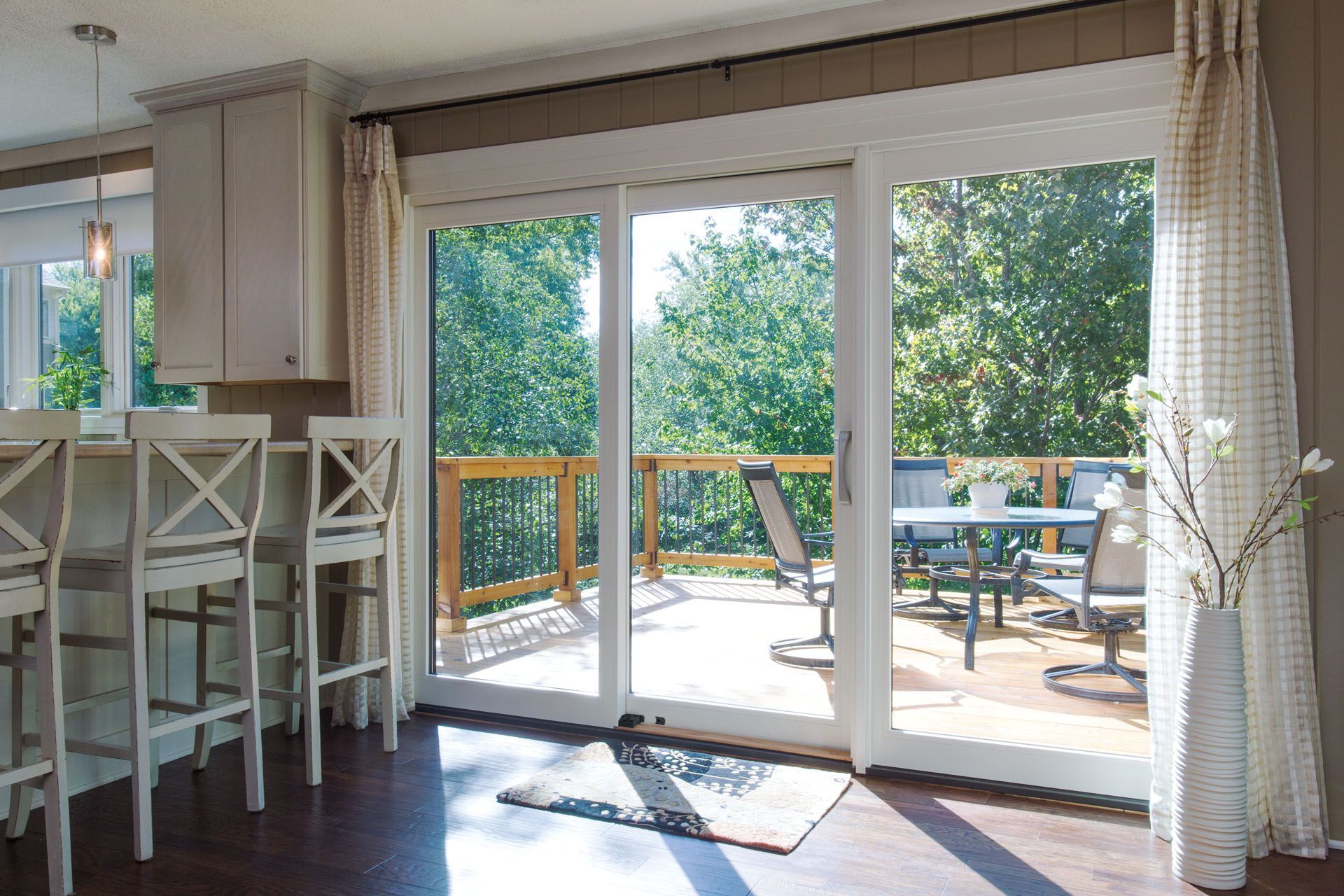 Bright kitchen with sliding glass door to a deck with table and chairs, trees visible.