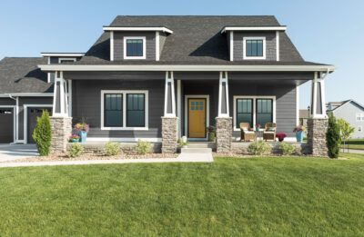 Gray house with yellow door, porch, stone pillars, green lawn, and blue sky.