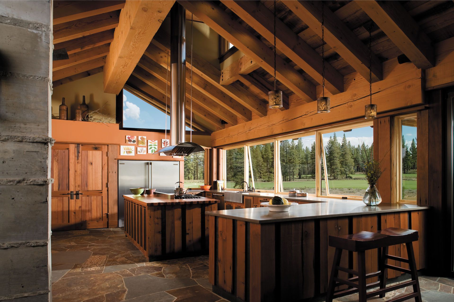 Rustic kitchen with wood beams, cabinetry, and large windows overlooking a forest.