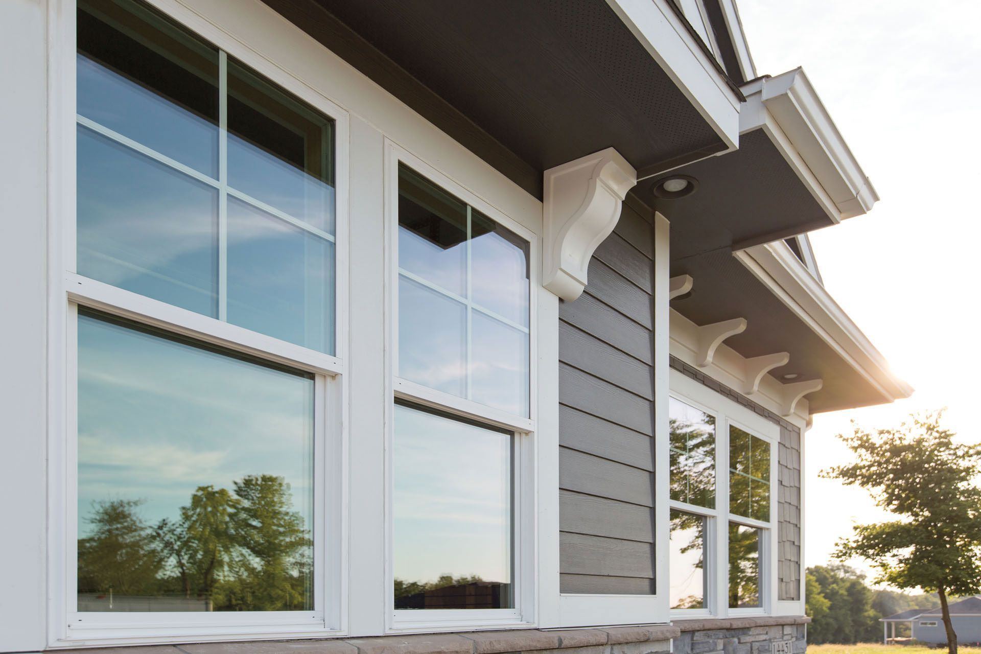 White-framed windows on a gray house with decorative supports; sky and trees reflected in the glass.