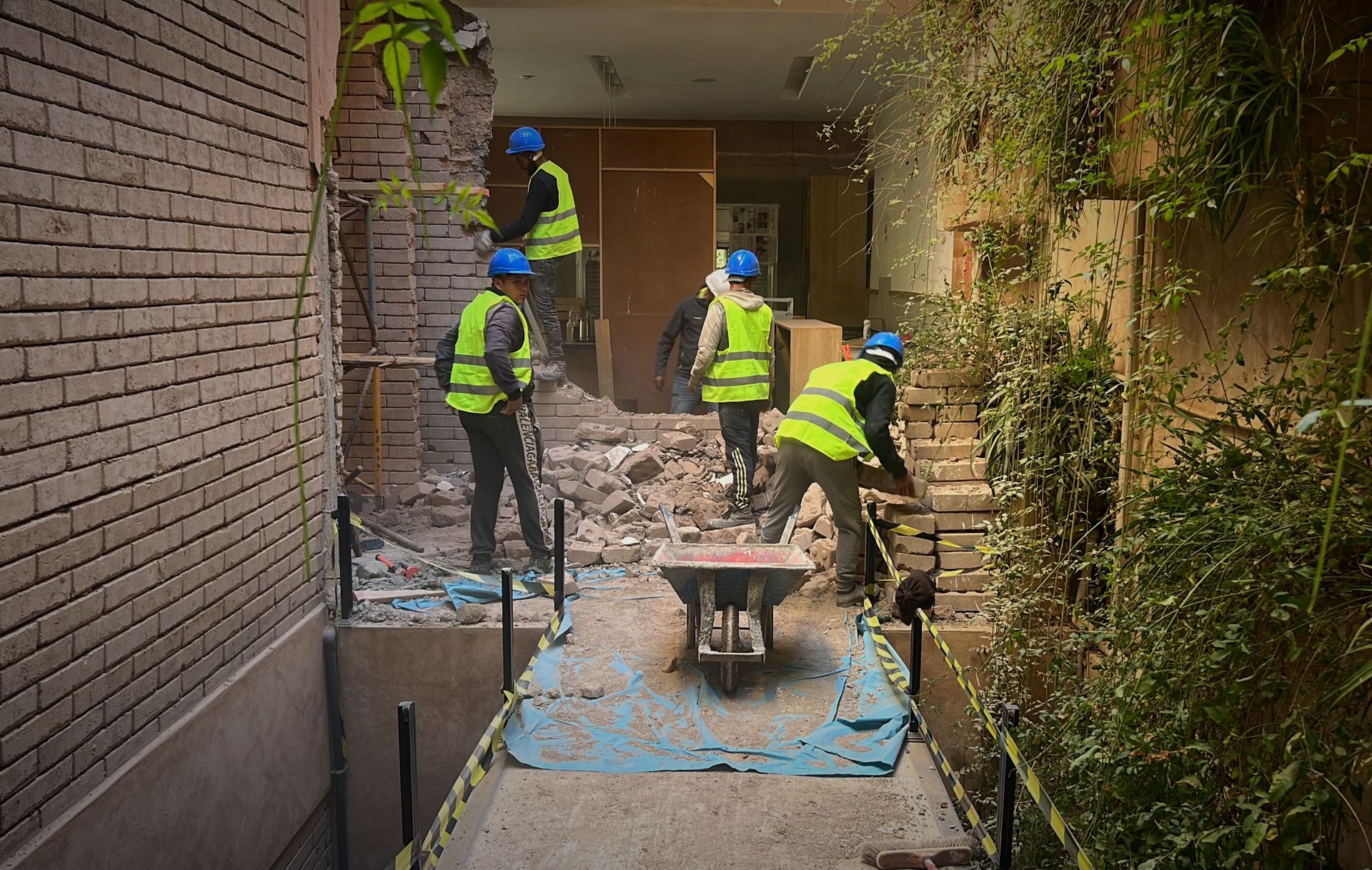 Des ouvriers du bâtiment démolissent un mur de briques dans un espace étroit, portant des gilets de sécurité et des casques.