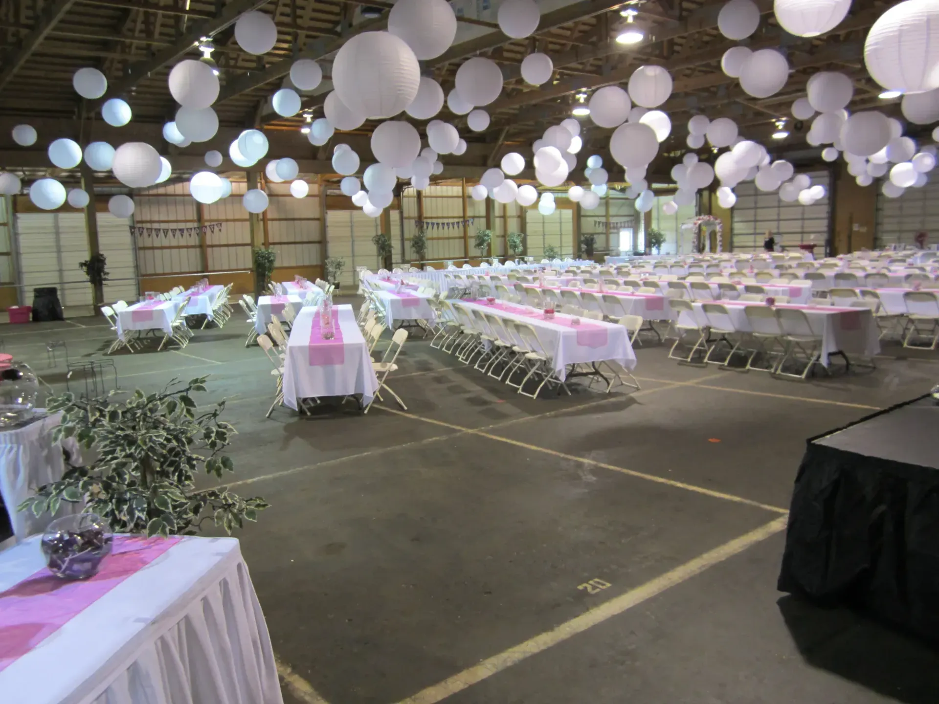 Large event hall decorated with white lanterns, tables with pink runners, and white chairs.