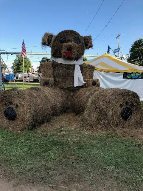 Hay bale teddy bear sculpture with red lips, white scarf, in grassy area.