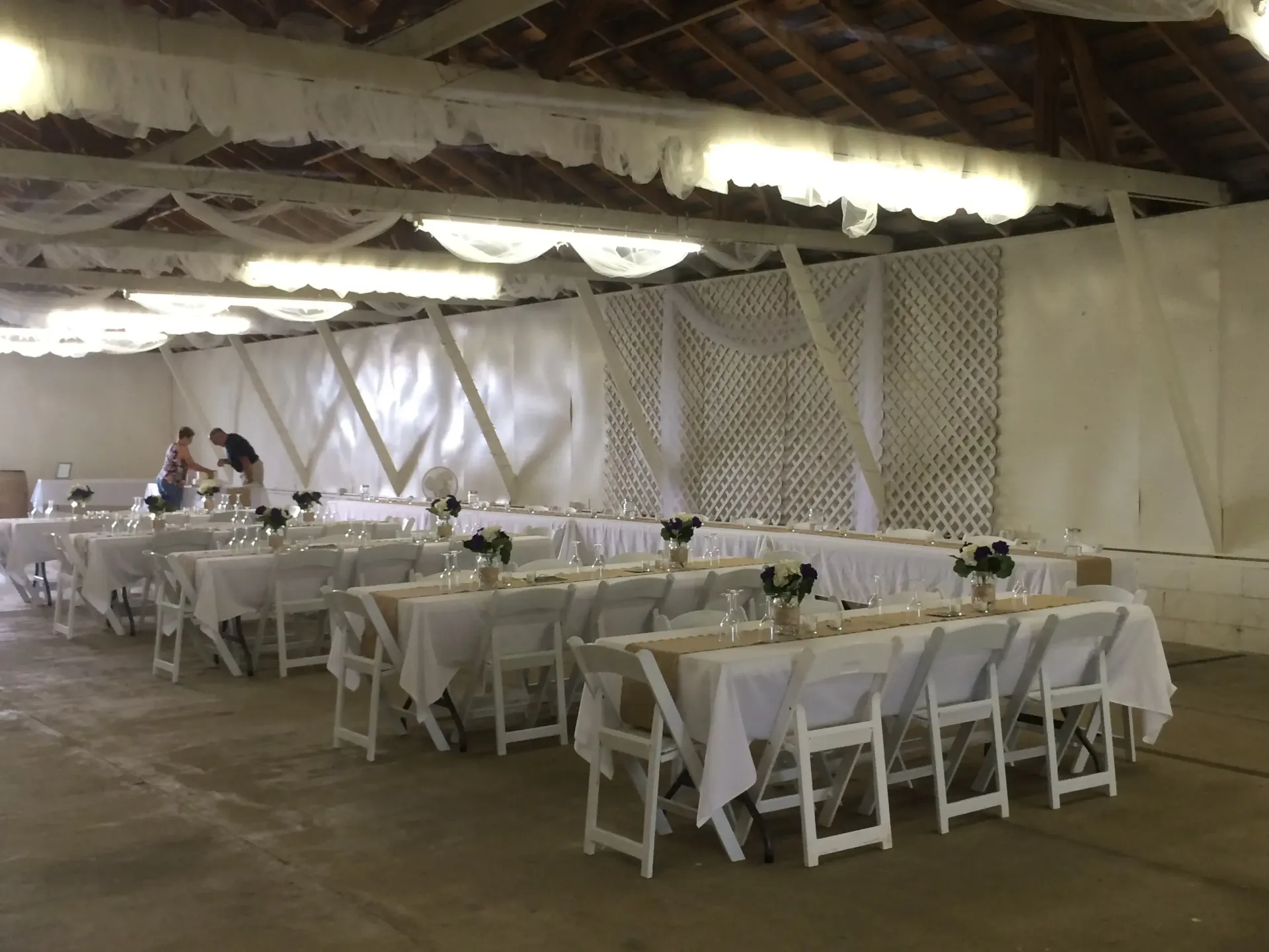 Long tables set for a gathering inside a white-walled building. White chairs and floral centerpieces are visible.
