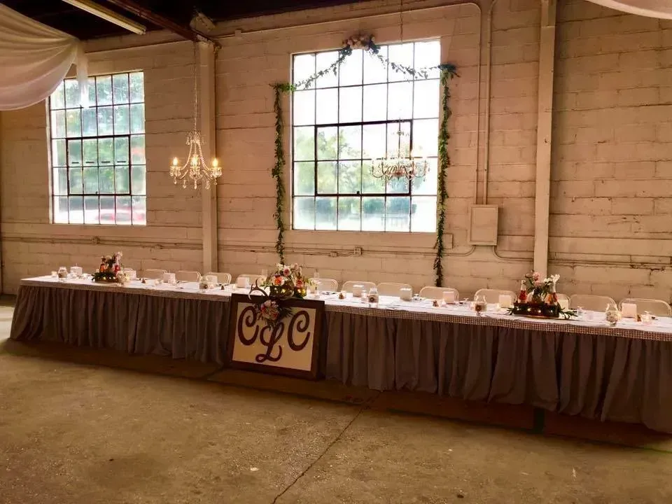Wedding reception head table with centerpieces, initials, and chandeliers in a venue with large windows.