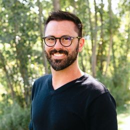 Man with glasses smiles, looking at the camera. He wears a black v-neck shirt, with a background of trees.