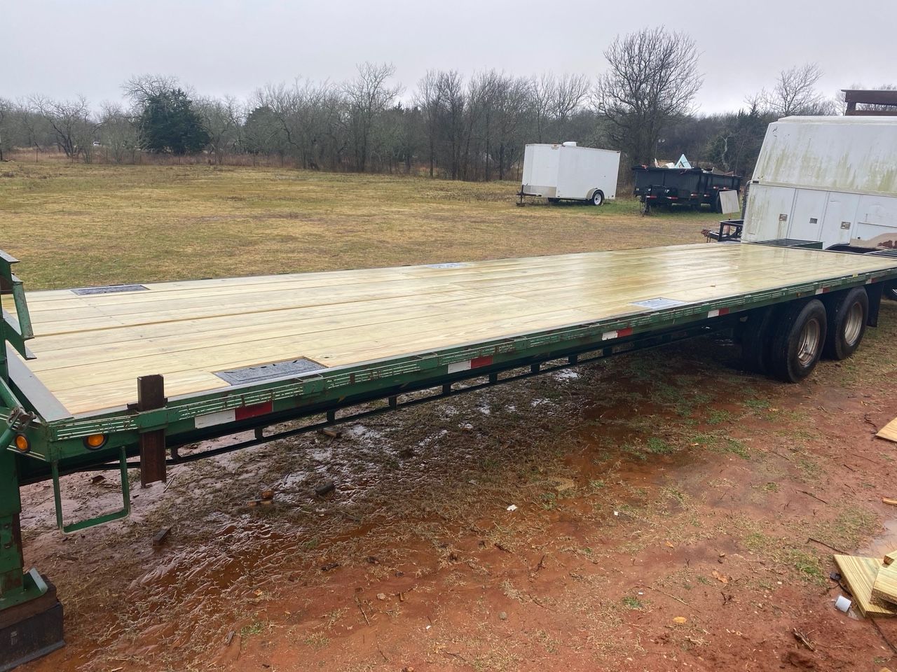 Flatbed trailer on a muddy, grassy field with a backdrop of trees and other trailers.