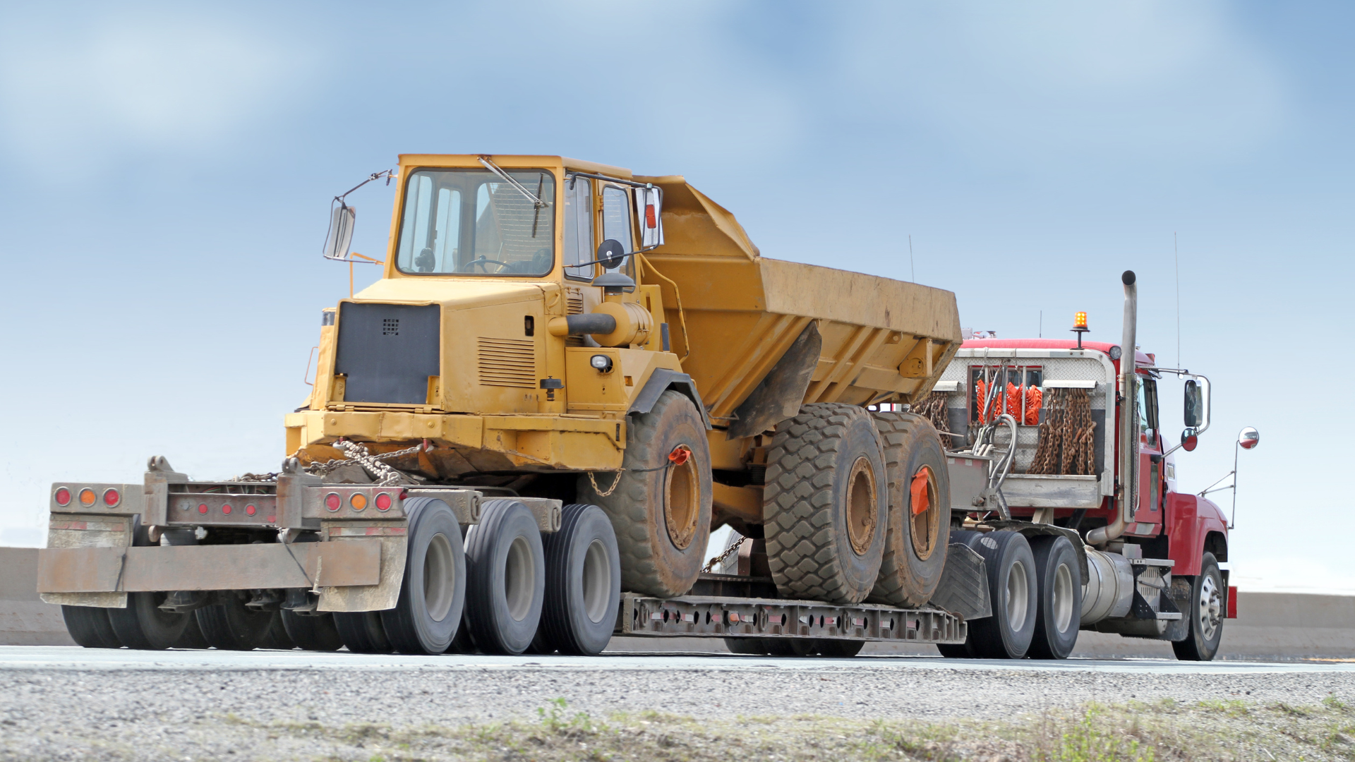 A red semi-truck carries a large yellow articulated dump truck on a flatbed trailer along a road against a blue sky.