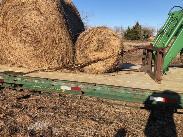Two hay bales on a trailer, one secured with a chain to a tractor's forks, set in a field.