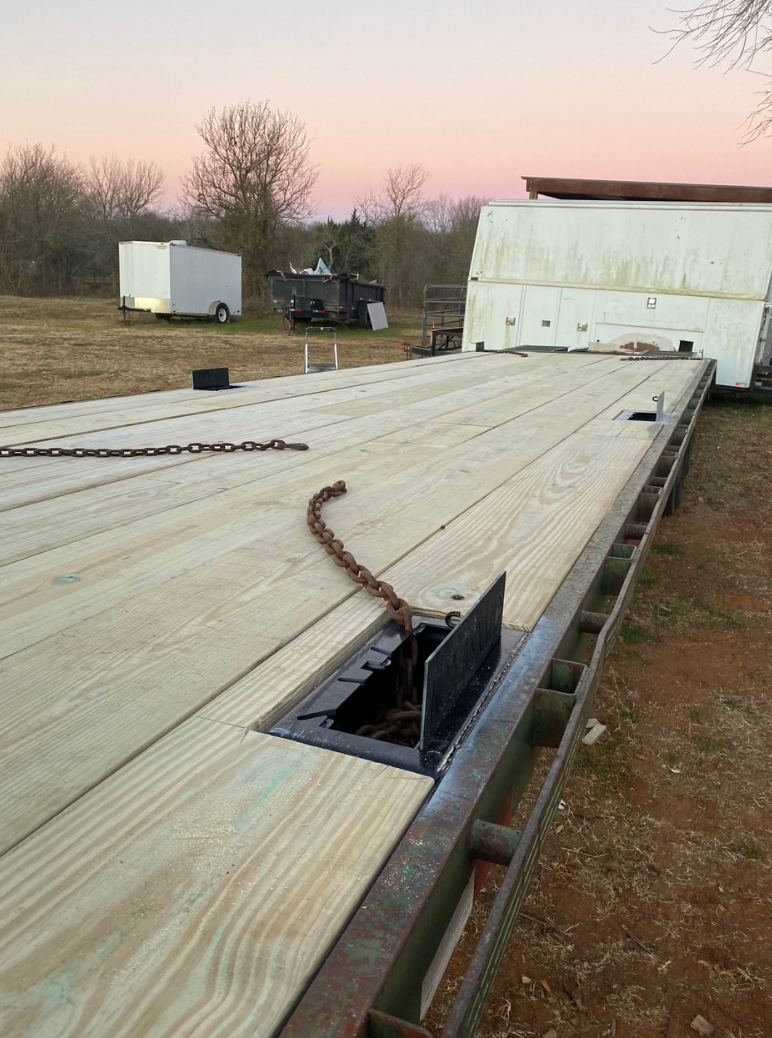 Flatbed trailer with wooden deck, chains, and tie-down points outdoors at dusk.