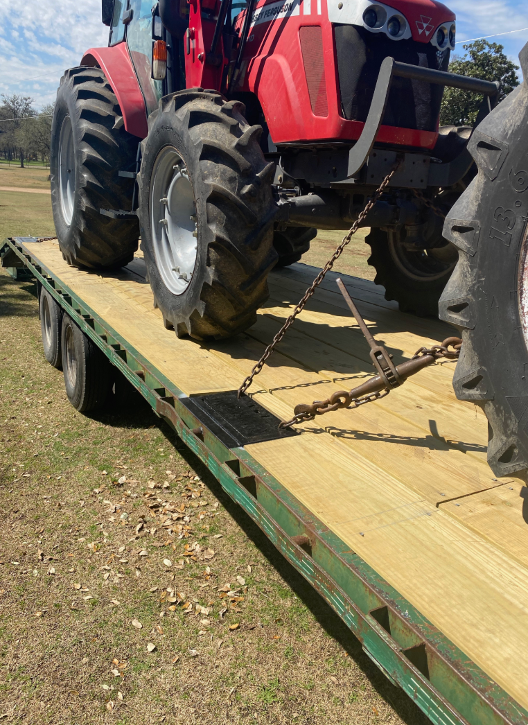 Red tractor on a wooden flatbed trailer, secured with chains, outdoors on grass.