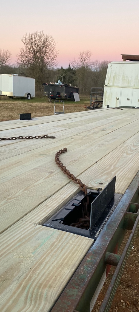 A flatbed trailer with wooden decking features a recessed chain storage box holding a chain at sunset in a rural field.