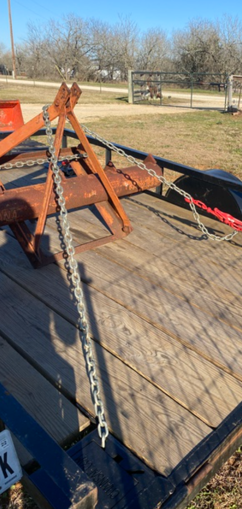 A metal A-frame implement secured with a chain and red load binder on the wooden bed of an open utility trailer.