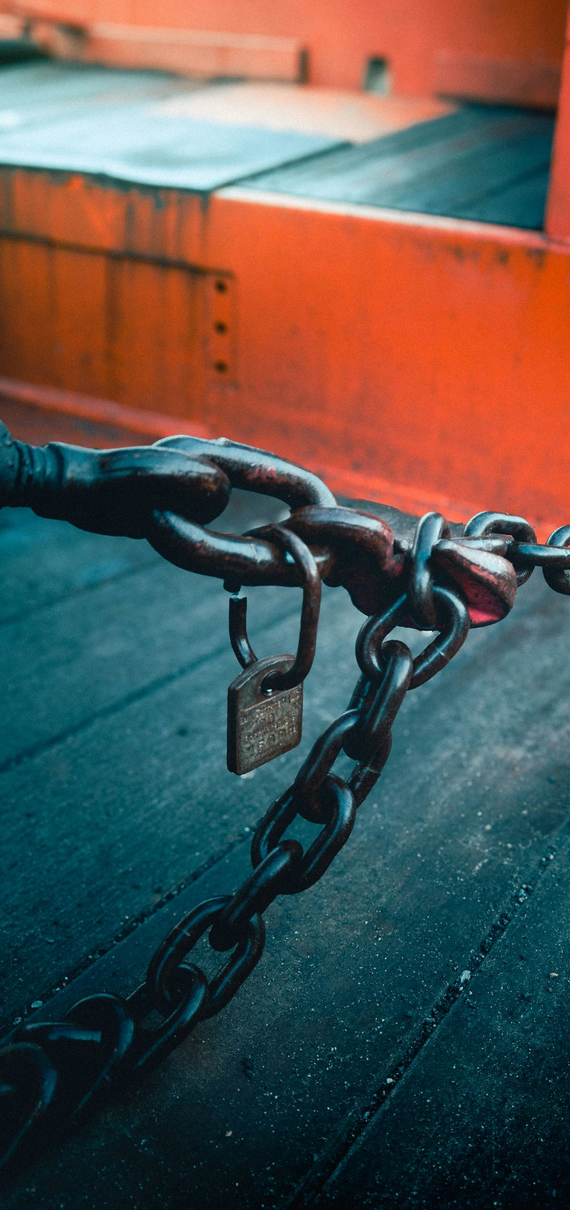 A close-up of a metal chain and padlock secured to an orange industrial structure against a dark, wooden background.