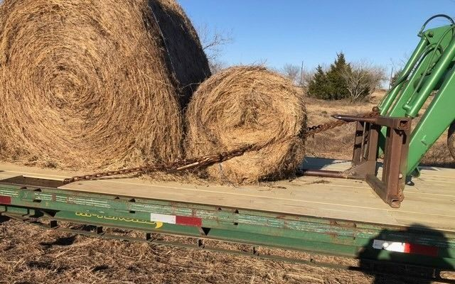 A tractor loader spear is positioned to lift a round hay bale on a flatbed trailer next to a larger bale.
