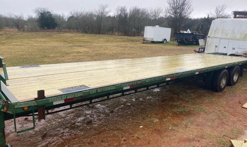A long, green flatbed utility trailer with new, light-colored wooden planks, parked on a muddy rural field.