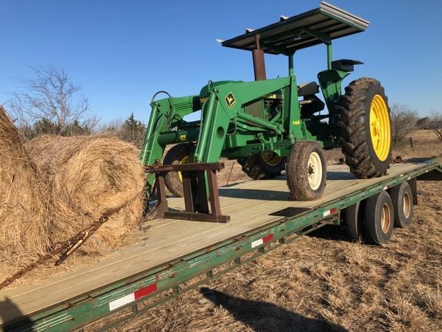 Green John Deere tractor with loader on a trailer, hay bales in the background, sunny day.