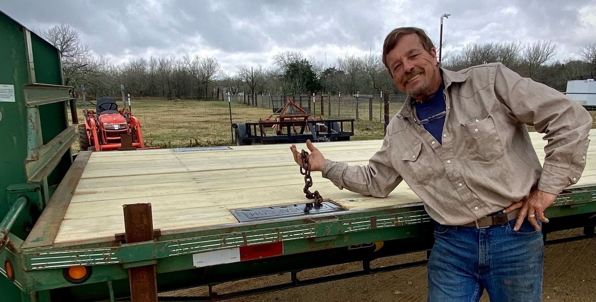 Man with chain on flatbed trailer; tractor in background, cloudy day.
