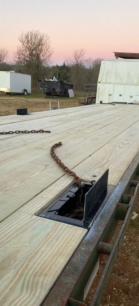 A flatbed trailer with a wooden deck and a recessed metal tie-down point in a grassy field at twilight.