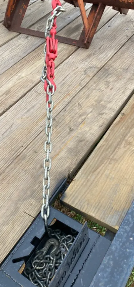 A metal chain with a red binder attached, connected to a steel tie-down point on a wooden trailer floor.