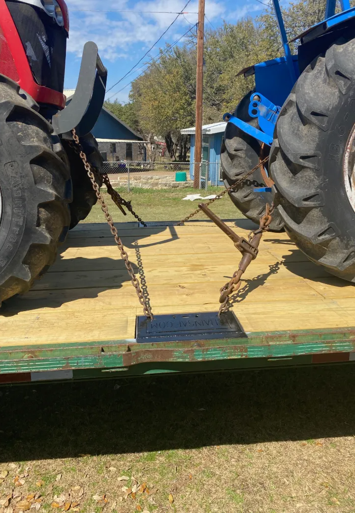 Two tractors are secured with chains to the bed of a flatbed trailer outdoors on a sunny day.
