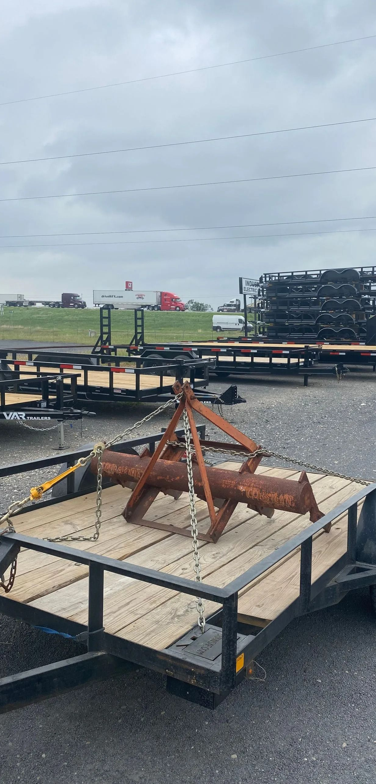 A utility trailer parked on gravel carries a rusty metal object secured by chains, with other trailers in the background.