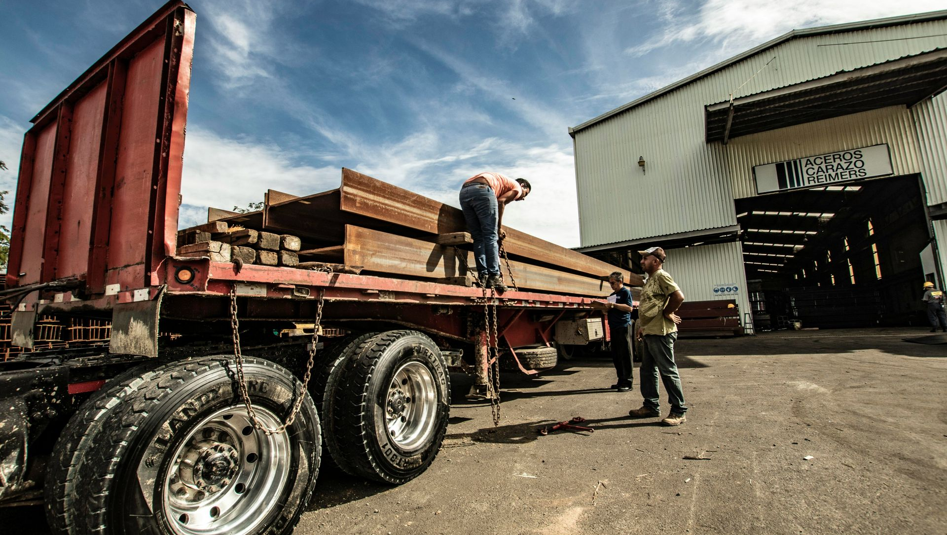 Two workers prepare a flatbed truck loaded with large steel beams outside a warehouse under a bright blue sky.