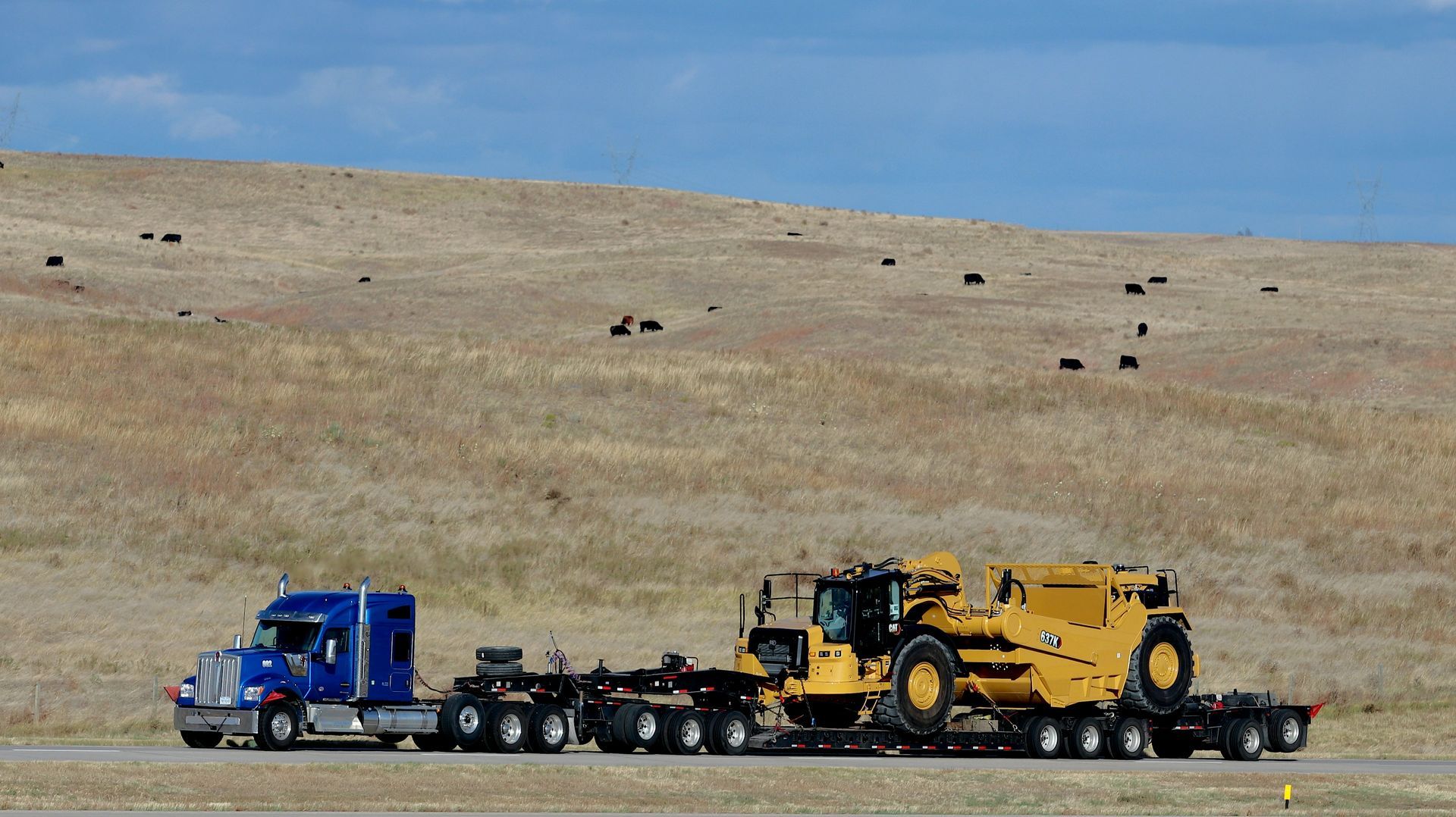 A blue semi-truck pulling a flatbed trailer carrying a yellow Caterpillar scraper on a road against a grassy, hilly backdrop.