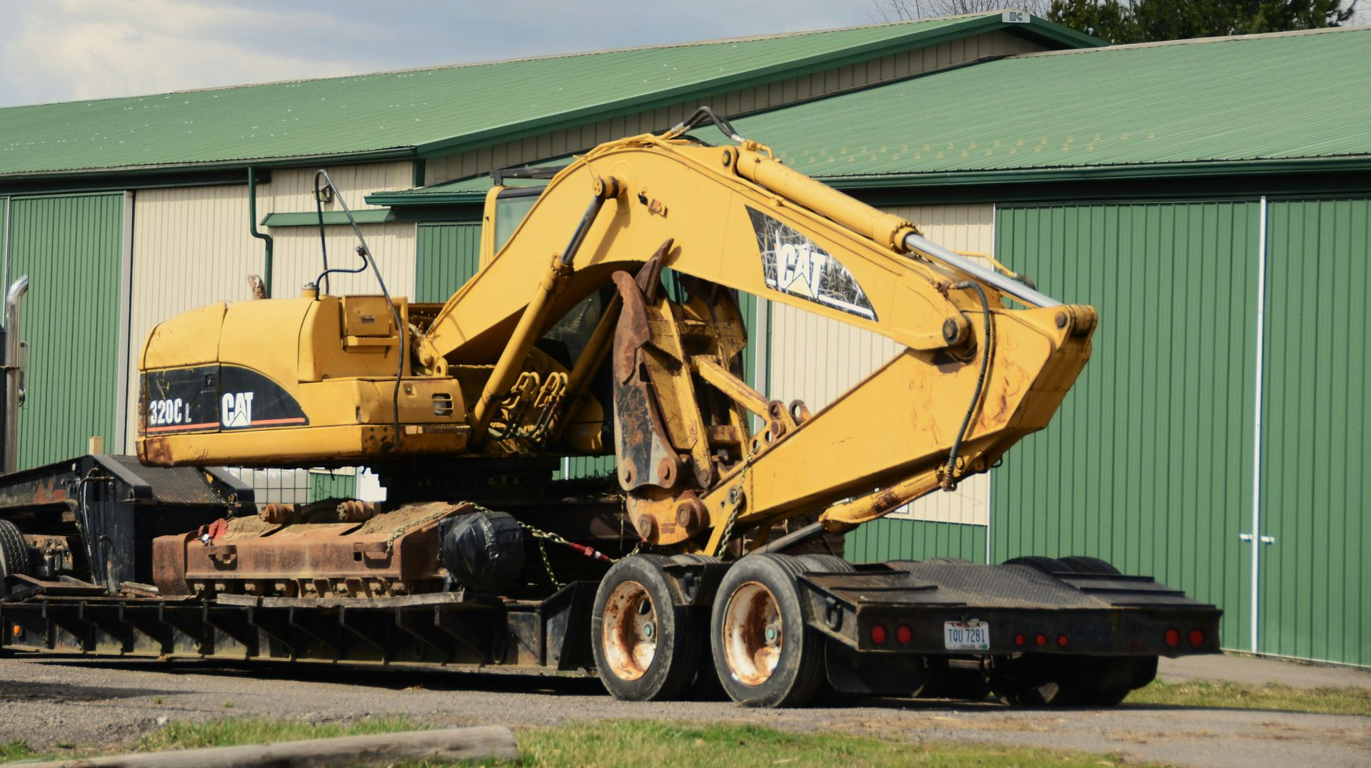 A yellow Caterpillar excavator is secured to a flatbed trailer parked in front of a building with a green roof.