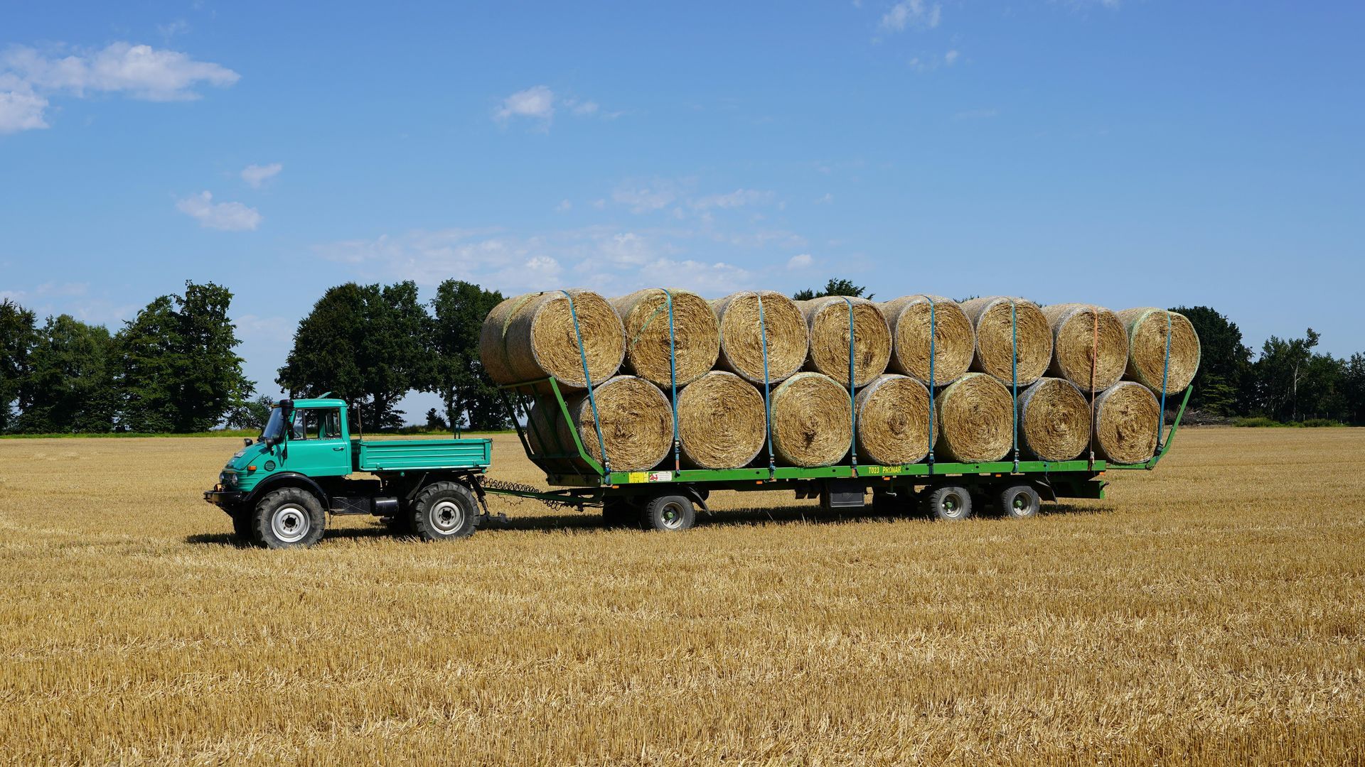 Green tractor hauling a flatbed trailer stacked with round hay bales across a harvested field under a blue sky